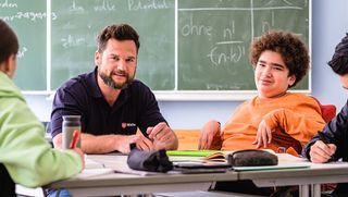 Malteser Schulbegleiter sitzt mit seinem Schüler und anderen Schülern in der Klasse. 