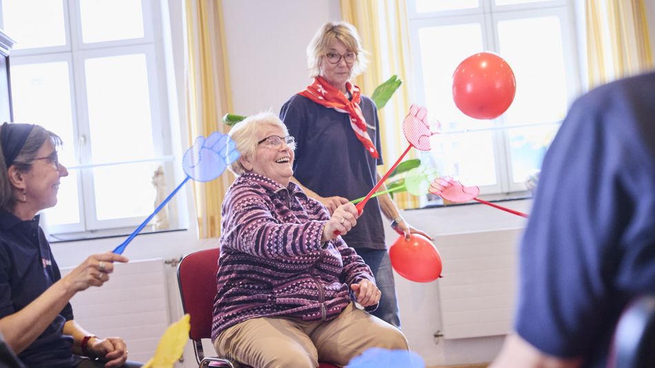 Mehrere Personen nehmen in einem hellen Gruppenraum an einer Aktivität mit bunten Luftballons teil. Eine Person sitzt auf einem Stuhl und bewegt mit einem Schläger einen roten Luftballon, während weitere Teilnehmende und Betreuungskräfte der Malteser die Übung begleiten.