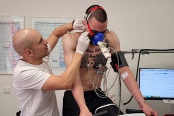 Ein Mann in Arztkleidung nimmt einer Person auf dem Fahrradergometer vom Ohr Blut ab.