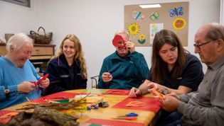 3 ältere Menschen sitzen zusammen mit 2 Malteserinnen an einem Tisch und schneiden zum Training der Feinmotorik etwas aus.