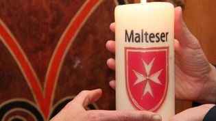 Hände, die eine brennende weiße Kerze mit dem Malteser Wappen halten. 