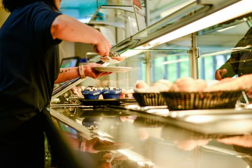Mitarbeitende Person reicht Essen an der Ausgabestation in einer Cafeteria.