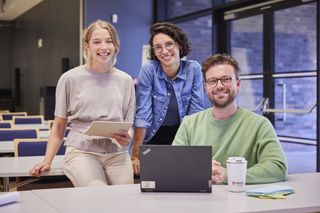 Drei junge Menschen – zwei Frauen und ein Mann – befinden sich in einem Schulungsraum der Malteser. Der Mann sitzt vor einem geöffneten Laptop am Tisch und die beiden Frauen sitzen links hinter ihm auf der Tischplatte. Alle drei lächeln in die Kamer