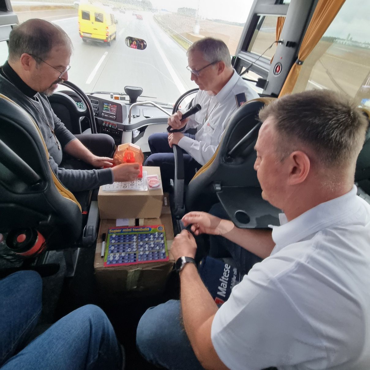 Drei Personen sitzen in einem Reisebus, zwei tragen weiße Hemden, einer schwarze Kleidung, auf dem Tisch zwischen ihnen liegen Gegenstände.