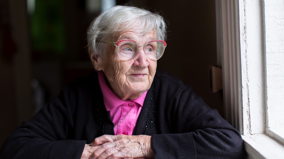 Eine alte Frau mit Brille schaut aus einem dunklen Raum aus einem Fenster und lächelt leicht.