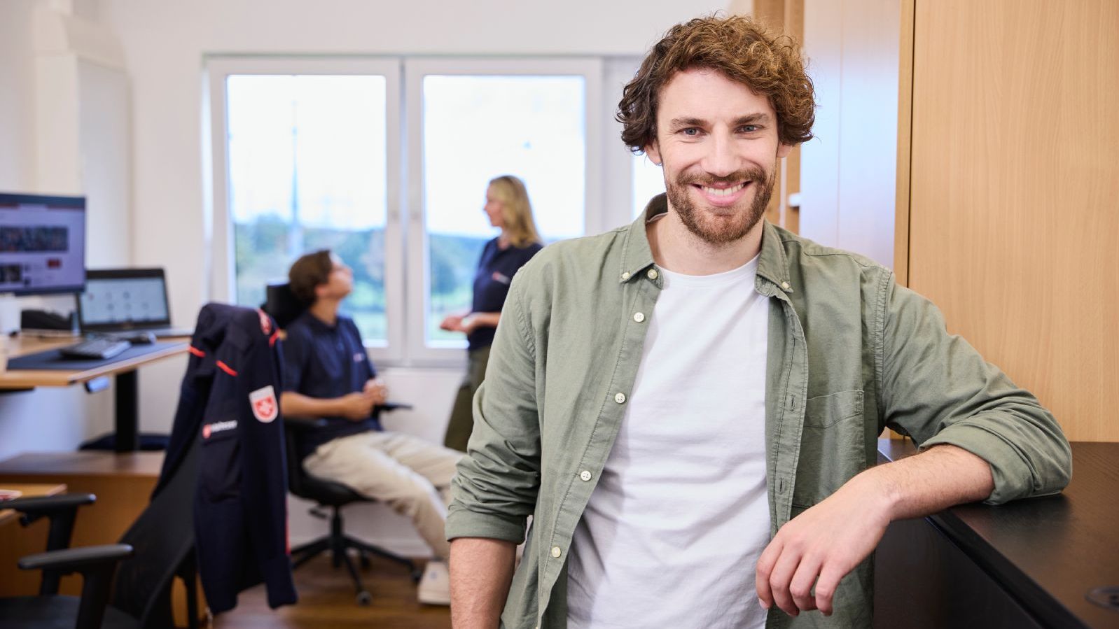 Mitarbeiter der Malteser steht im Vordergrund in einem Büro, im Hintergrund unterhalten sich zwei Mitarbeitende vor einem Arbeitsplatz