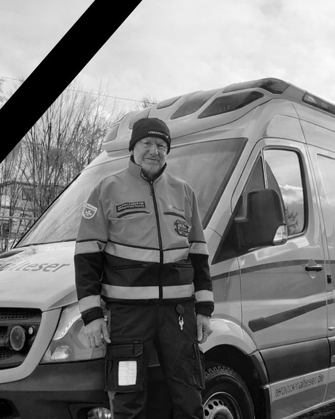 Eine Person in Einsatzkleidung der Malteser steht vor einem Einsatzfahrzeug. Das Bild ist in Schwarz-Weiß gehalten, oben links befindet sich ein Trauerband. Es handelt sich um René Kluge.