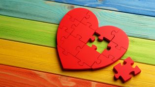 Rotes Herz aus Puzzleteilen, ein Teil fehlt und liegt daneben, auf buntem Holzuntergrund in Regenbogenfarben.