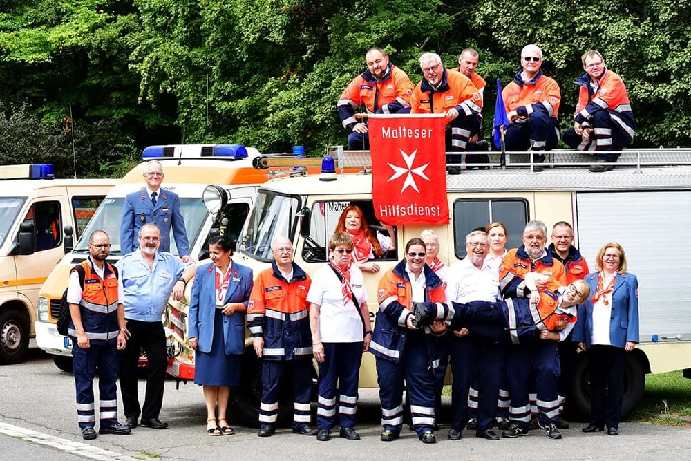 Gruppenfoto von Frauen und Männern in Malteser Dienstbekleidung vor und auf Dienstfahrzeugen. 