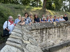 Auf dem Foto sind mehrere Personen zu sehen, welche an einer Mauer stehen. Im Hintergrund sieht man Hügel und Bäume