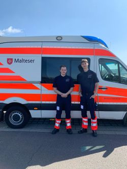 Zwei junge Mitarbeitende des Malteser Hilfsdienst e.V. stehen vor einem orange‑weiß markierten Rettungswagen. Sie tragen dunkle Polohemden mit Logo und schauen direkt in die Kamera — das Bild vermittelt Einsatz‑ und Helferbereitschaft.
