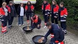 Mehrere Personen in Malteser Kleidung stehen um zwei Feuerschalen, an denen zwei Personen ein Feuer entzünden