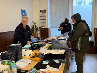 Ein Frau in Malteser Kleidung steht hinter einem Tisch mit Stapeln von verschiedener warmer Kleidung. Vor dem Tisch steht ein Mann mit Kapuzenjacke. 