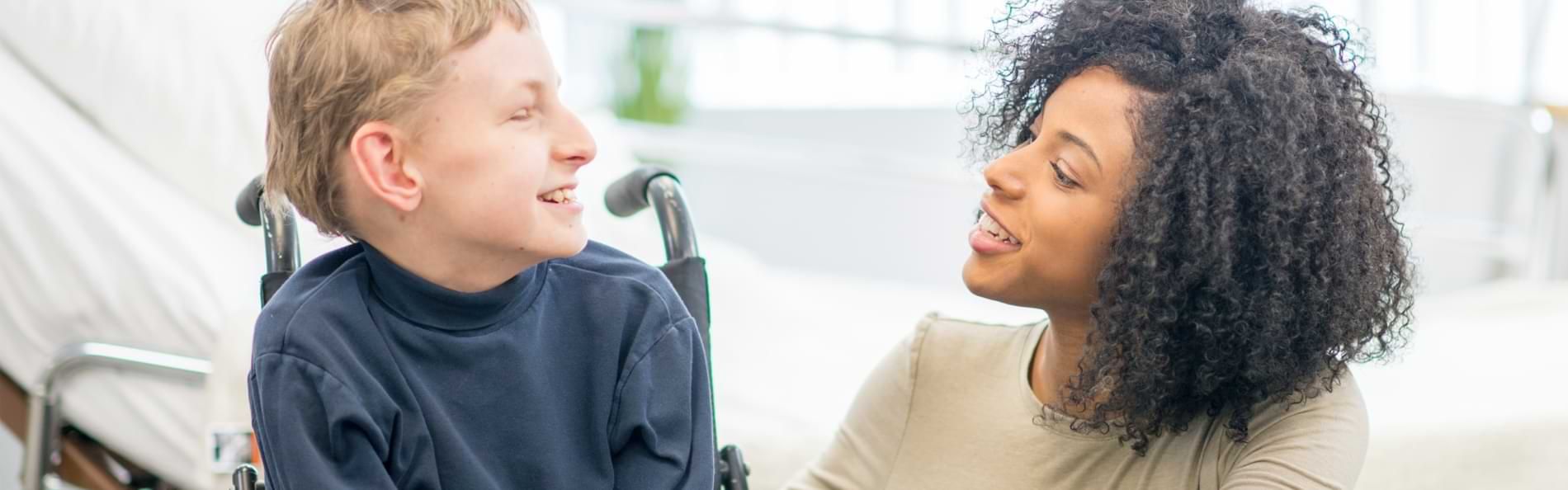 Ein Junge im Rollstuhl und eine knieende junge Frau in einem Krankenzimmer.