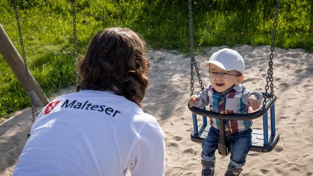 Eine Malteser-Pflegekraft schaukelt einen Jungen auf einem Spielplatz.