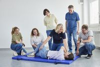 Eine Frau in Malteser-Kleidung dreht einen Mann in Seitenlage, mehrere Menschen schauen zu
