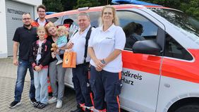 Eine Familie und Rettungskräfte stehen neben einem Notarztauto. Alle sind entspannt.