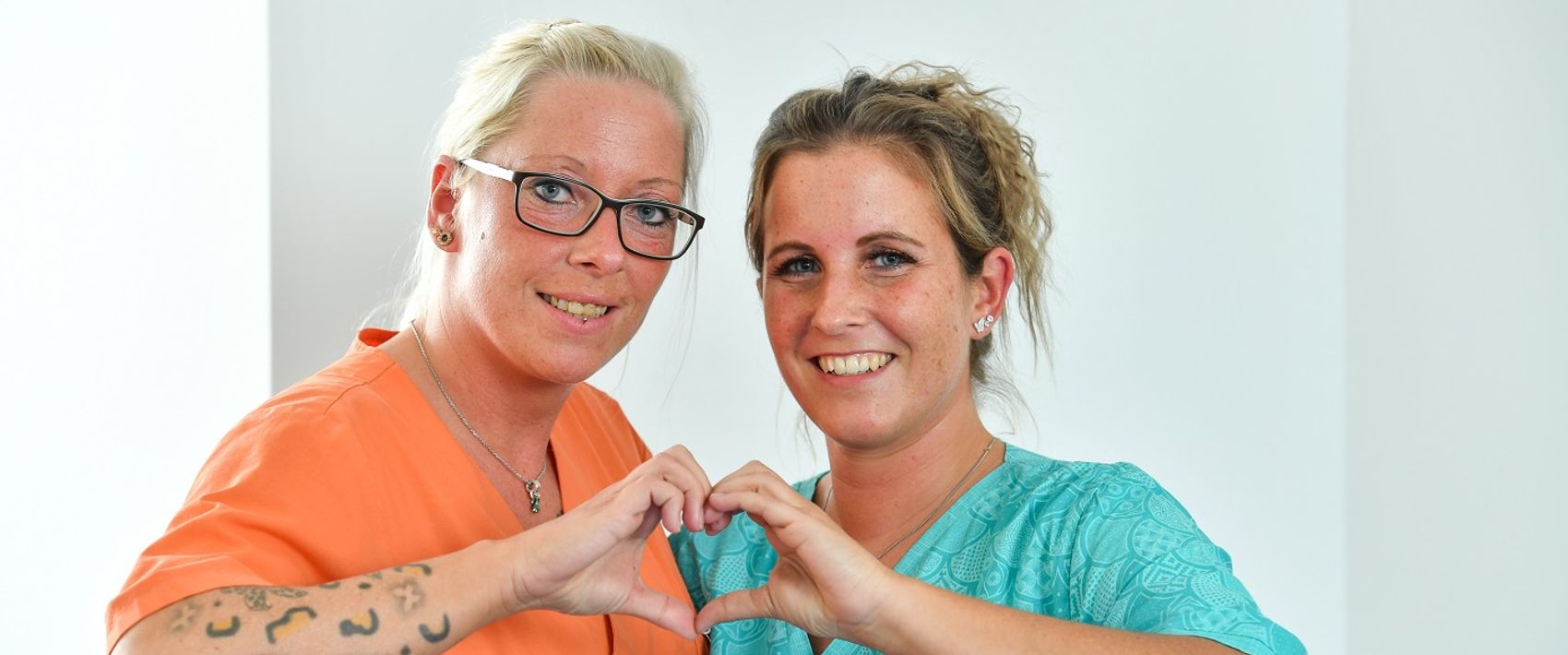Zwei Frauen formen gemeinsam mit den Händen ein Herz und lächeln dabei in die Kamera