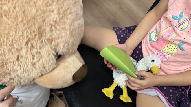 Ein Kind hält eine Stoffmöve mit "Papierschiene" in den Händen, beobachtet von einem Teddybären.