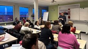 Menschen sitzen an Tischen und lauschen einem Dozenten