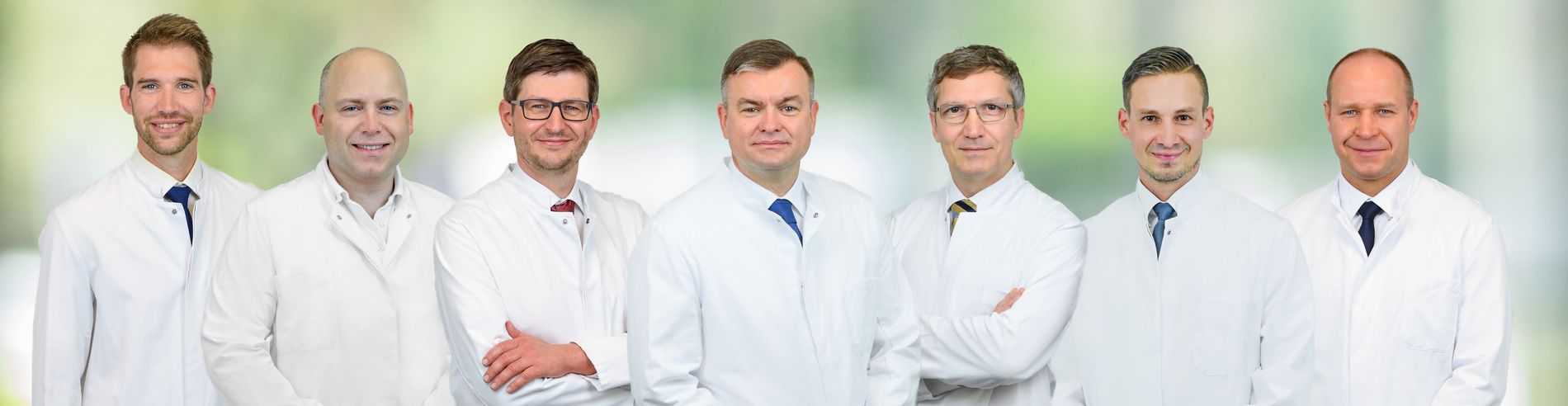 Sieben Ärztinnen und Ärzte in weißen Kitteln vor unscharfem Hintergrund, stehend nebeneinander und freundlich blickend.