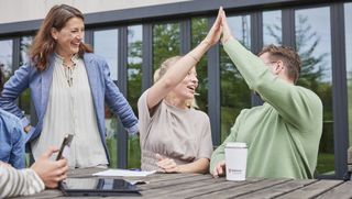 Zwei junge Menschen – links eine Frau, rechts ein Mann – sitzen an einem Outdoor-Holztisch vor einem Bürogebäude und klatschen erfreut jeweils mit einer Hand mittig oberhalb ihrer Köpfe ab. Ein weitere Frau steht links daneben und lacht mit den beiden.