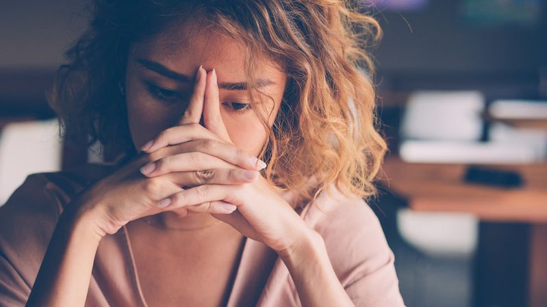 Eine junge Frau in einem Büro mit müdem Geschichtsausdruck und den Händen vor dem Gesicht