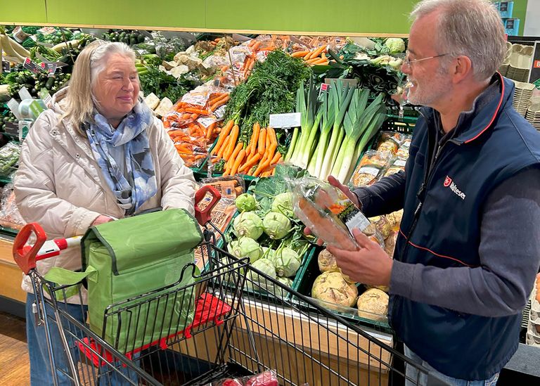 Alte Frau mit Einkaufswagen und Malteser Ehrenamtler vor einer Gemüseauslage im Supermarkt.