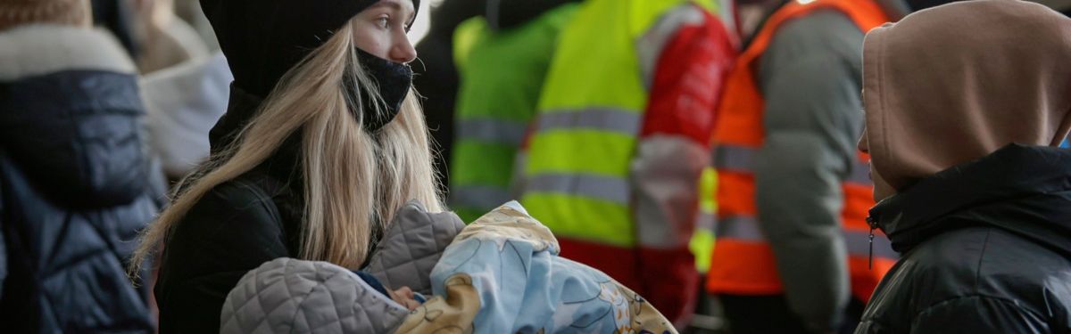 Schwarz gekleidete Frau mit einem Kleinkind im Arm. Im Hintergrund Menschen mit Warnwesten.