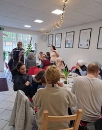 Menschen verschiedener Altersgruppen sitzen an mehreren Tischen in einem hellen Raum und kommen bei Kaffee, Kuchen und Spielen ins Gespräch. Der Treffpunkt ist festlich dekoriert, im Hintergrund sind Ehrenamtliche der Malteser zu sehen.