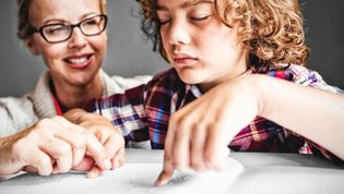 Frau sitzt hinter einem Jungen, der in Blindenschrift liest.