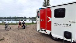 Das MalteserMobil ist zu sehen, ein Auto für die Obdachlosenhilfe. Zwei Frauen sitzen daneben auf einer Bank. Im Hintergrund ist Wasser zu sehen.