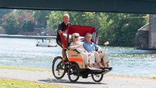 Zwei Senioren werden mit einer Malteser Rikscha an einem Fluss entlang gefahren.