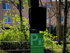 Ladesäule für Elektrofahrzeuge mit grünem Hinweisschild vor einem Gebäude im Grünen.