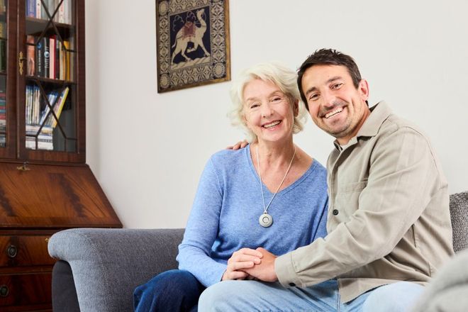 Eine ältere Dame mit Hausnotruf-Knopf sitzt mit ihrem Sohn auf dem Sofa und lacht