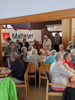 Zahlreiche Menschen sitzen an Tischen und besuchen das Buffet vor einem Banner der Malteser