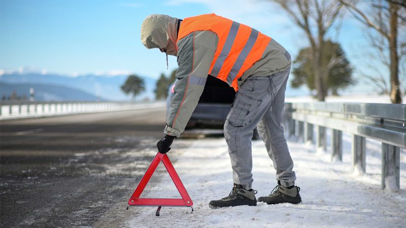 Eine Person in Winterbekleidung und einer Warnweste stellt ein Warndreieck im Schnee auf einer Straße auf.