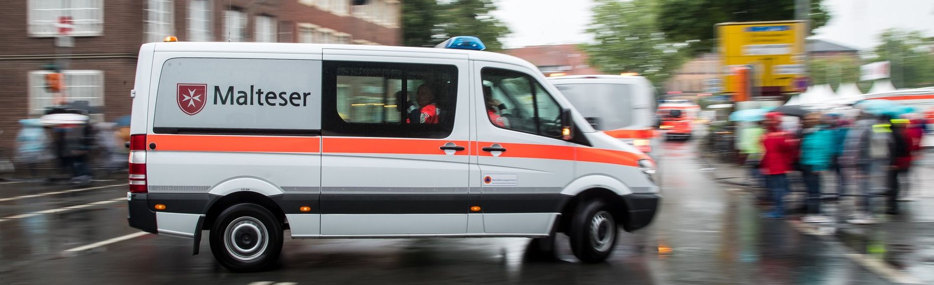 Ein Malteser Einsatzfahrzeug fährt im Vordergrund. Der Hintergrund mit Menschen und Fahrzeugen ist unscharf und verschwommen.