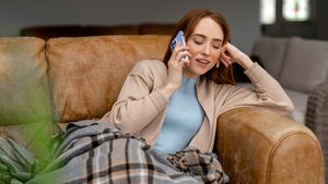 Eine junge Frau sitzt mit einer Decke gemütlich auf einem Sofa und telefoniert mit einem Smartphone