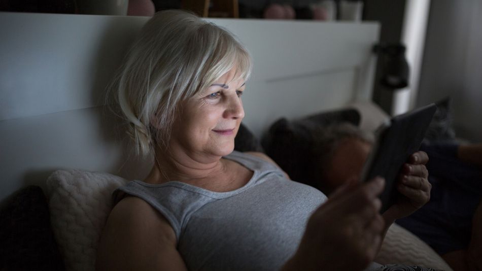 Ältere, lächelnde Frau liest im Bett mit einem E-Book-Reader.