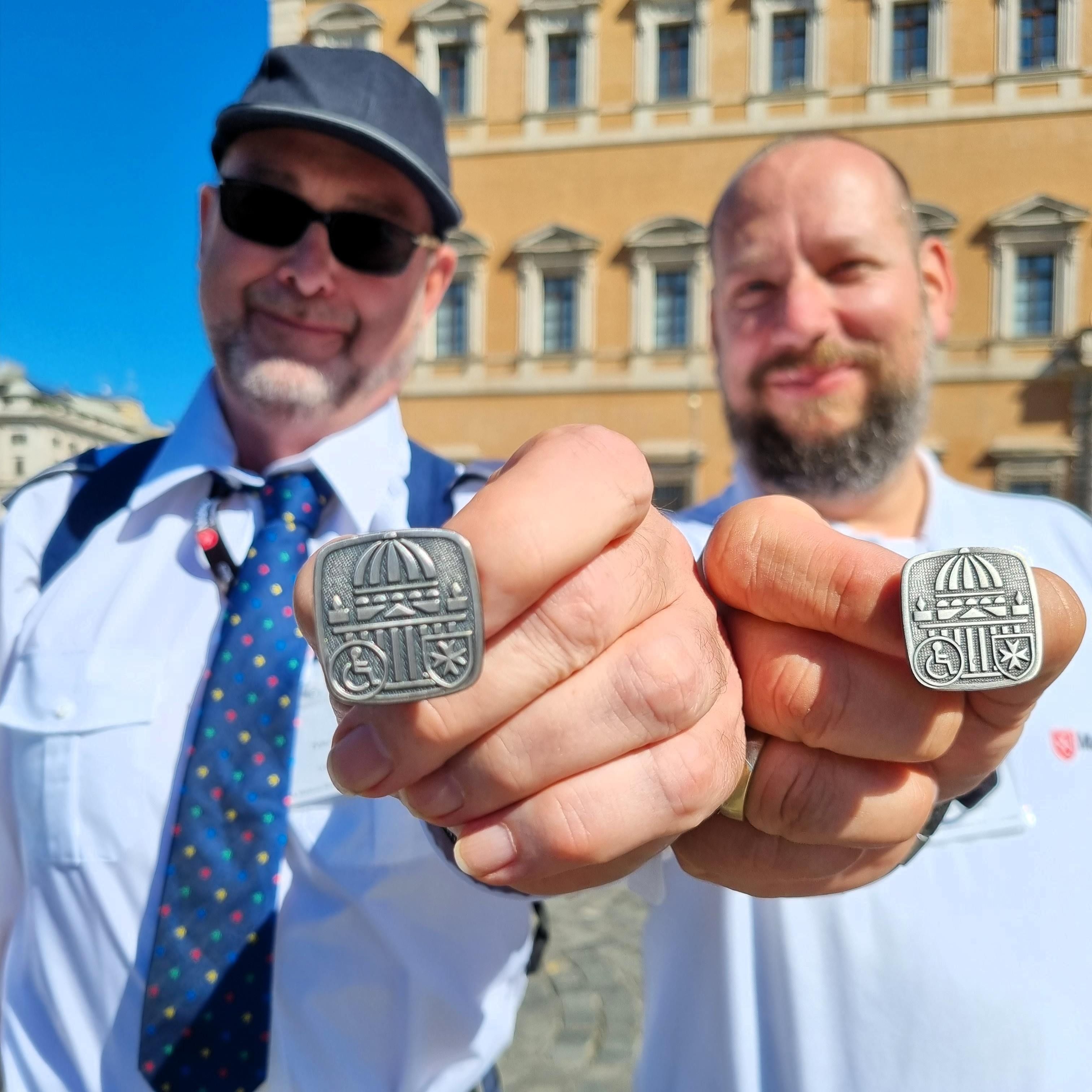 Zwei Männer zeigen jeweils eine quadratische Romwallfahrt-Plakette mit erhabener Schrift in die Kamera, Gebäude im Hintergrund.