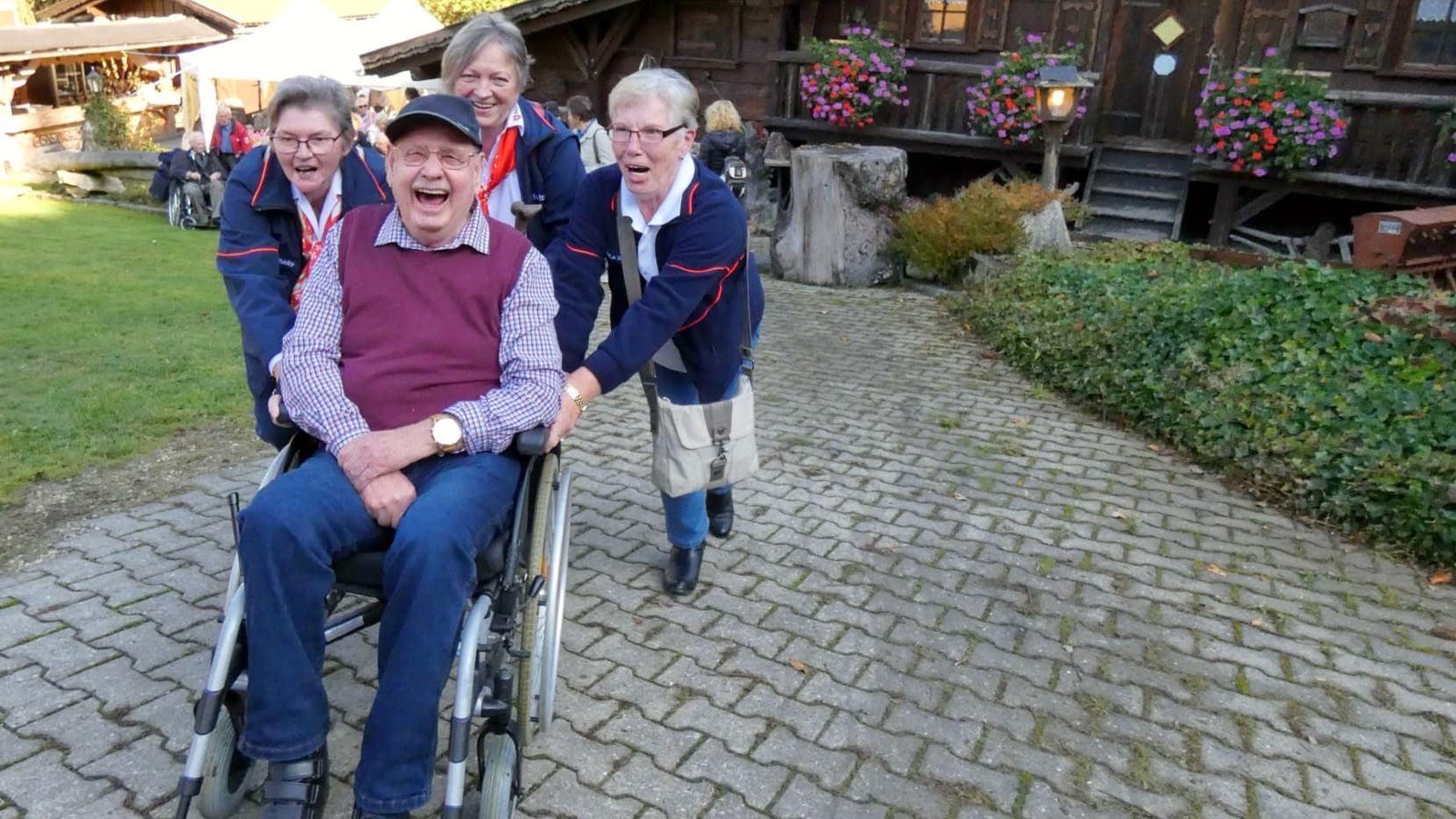 Lachender, älterer Rollstuhlfahrer wird von drei Frauen angeschoben.