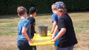 Vier Kinder tragen eine rote Rettungsbare aus Kunststoff. Darauf steht ein Eimer mit Wasser, welchen sie als Geschicklichkeitstest von A nach B tragen müssen.