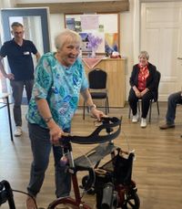 Eine Seniorin im blau-bunten T-Shirt probiert lachend einen Rollatur aus. Im Hintergrund rechts sitzt einen Malteser Mitarbeiterin und links steht ein Mitarbeiter des Sanitätshauses.
