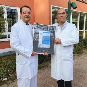 Zwei Ärzte in weißem Kittel stehen draußen und halten gemeinsam eine Urkunde mit einer Auszeichnung hoch.