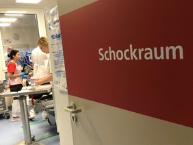 Blick durch geöffnete Tür mit Schild „Schockraum“ auf medizinisches Personal bei der Versorgung eines Patienten.