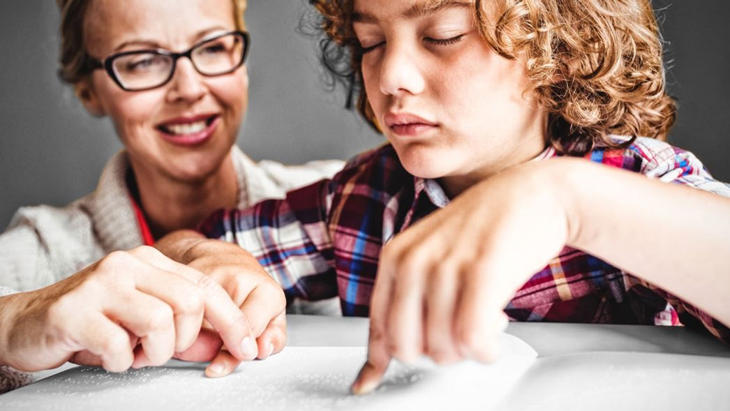 Frau sitzt hinter einem Jungen, der in Blindenschrift liest.