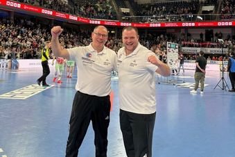 Zwei Männer in Sportkleidung auf einem Handballfeld, im Hintergrund Zuschauer und Hallenbeleuchtung.