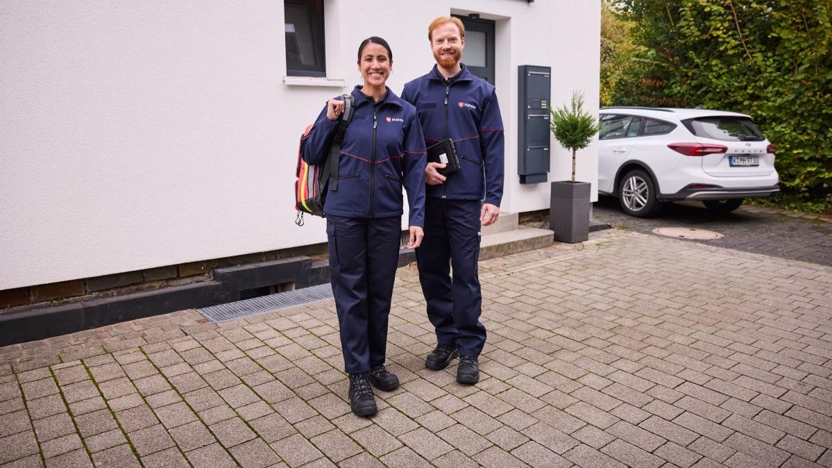 Zwei Mitarbeiter des Malteser Hausnotruf stehen vor einem Haus. Im Hintergrund ein Auto.