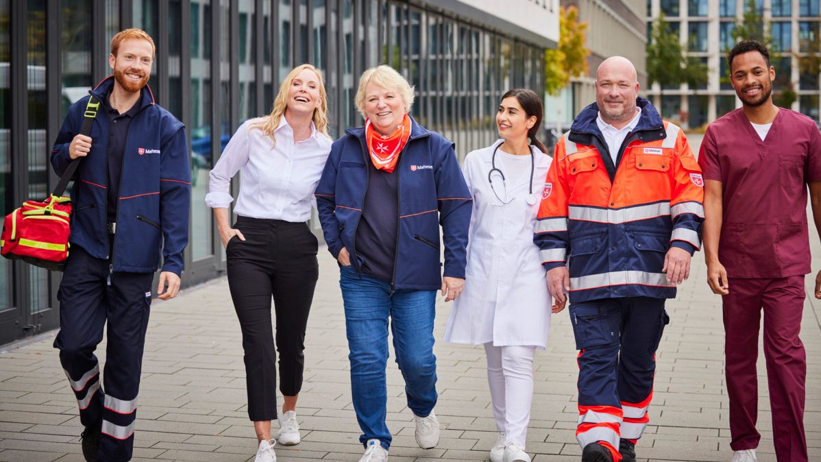 Sechs Malteser Mitarbeitende, die lachend in einer Reihe nebeneinanderher gehen und über unsere Stellenangebote für Quereinsteiger ihren Arbeitsplatz bei uns gefunden haben. Die Gruppe bildet die verschiedenen Jobs für (zum Beispiel) Quereinsteiger bei den Maltesern ab.